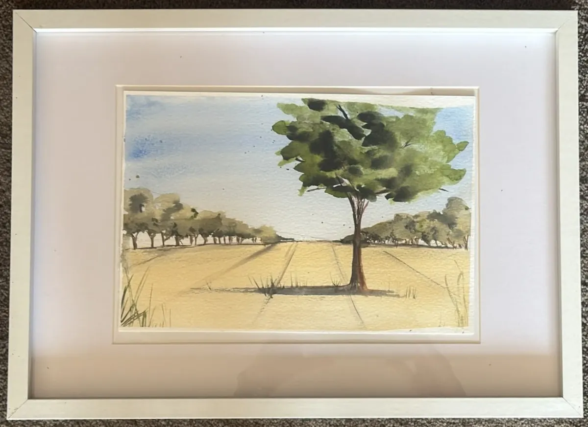 Field View: watercolour of a lone tree in a golden field with a line of trees beyond, mounted in a white frame.