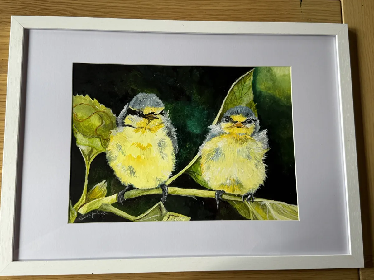 Fledglings: acrylic painting of two fluffy baby blue tits perched on a leafy branch, mounted in a white frame.