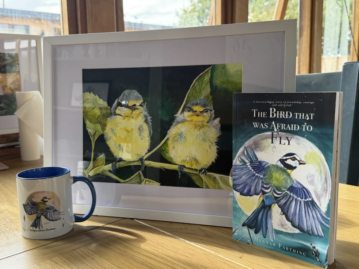 The framed Fledglings painting displayed with The Bird That Was Afraid to Fly book and mug.