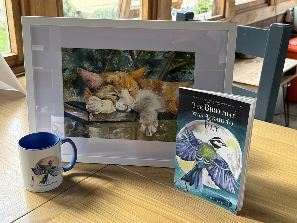 The framed Thistle painting displayed with The Bird That Was Afraid to Fly book and mug.