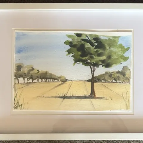 Field View: watercolour of a lone tree in a golden field with a line of trees beyond, mounted in a white frame.