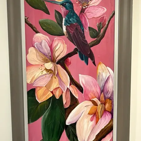 Hummingbird in Magnolia: acrylic painting of a green hummingbird among pink magnolia flowers, in a tall grey frame.